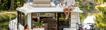 Un couple détendu profite de la boat life sur la terrasse d'un bateau habitable tout confort navigant sur un canal ombragé, avec des vélos à bord.